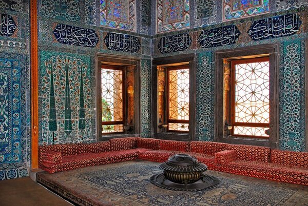 Sala de Topkapi decorada con azulejos azules de İznik, divanes rojos y un brasero central de latón; luz natural con celosías y frisos caligráficos.